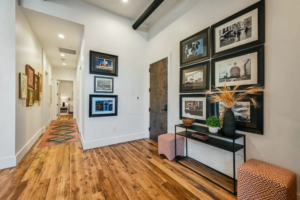 This image features a hallway with a gallery wall adorned with framed photos, a decorative console table, and textured ottomans. The warm wood flooring and artistic décor create a sense of connection to the city’s creative energy and urban lifestyle.