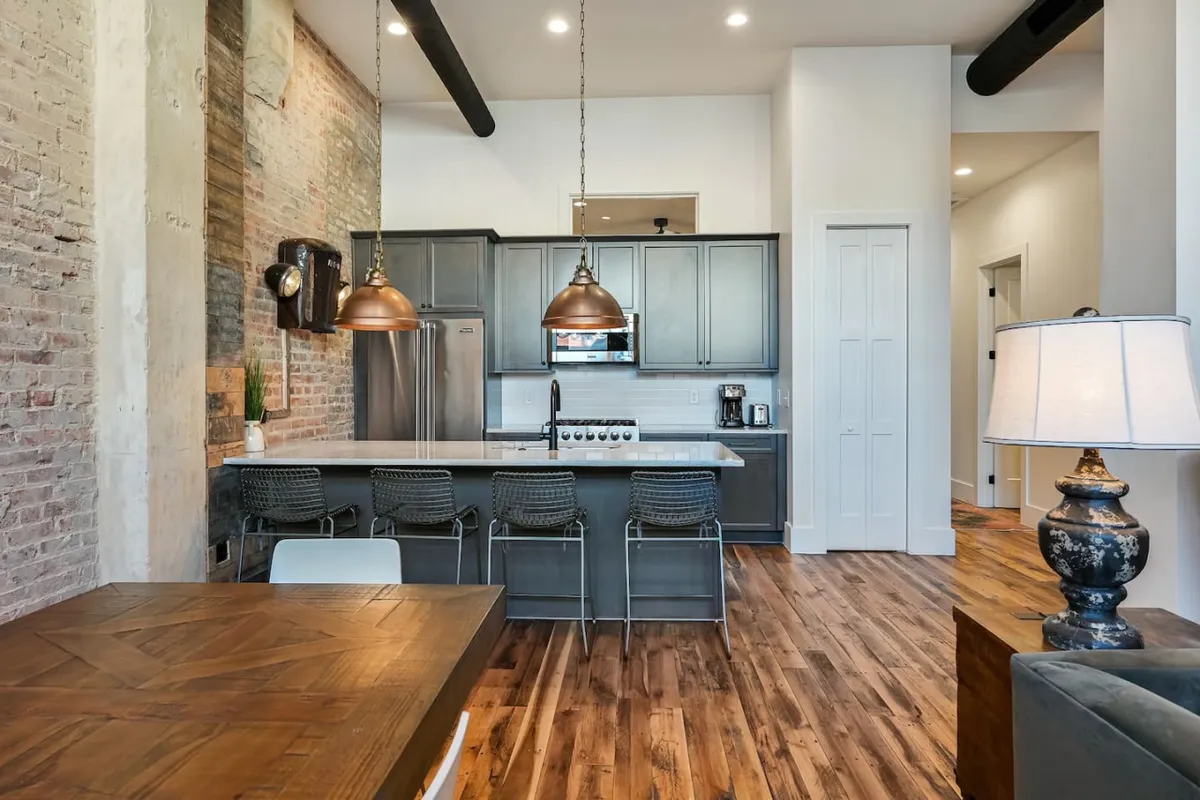 This image captures a modern kitchen with a central island, copper pendant lights, an exposed brick wall, a wood dining table, and stainless steel appliances. The layout supports socializing and group dining, with visual cues of a welcoming environment for cooking and celebrating.