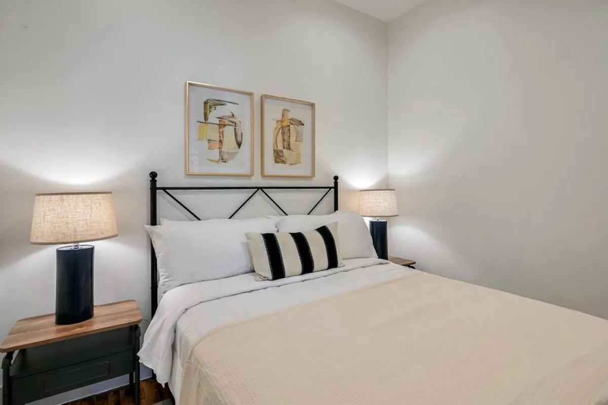 A serene bedroom with a queen bed dressed in soft linens and decorative pillows, complemented by two bedside lamps and modern artwork. Gentle natural light fills the space, offering a tranquil environment ideal for relaxation or quiet reading.