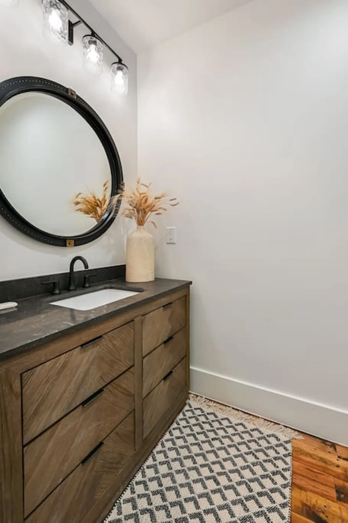 A modern half bath featuring a large round mirror, wooden vanity, decorative vase with dried plants, and a textured rug. The serene atmosphere and quality materials evoke a boutique hotel feel, emphasizing comfort and style.