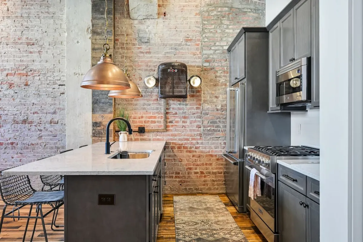 This image displays a stylish kitchen featuring copper pendant lights, a marble countertop, exposed brick wall, and modern appliances. A woven rug adds warmth to the space, and the overall ambiance is contemporary and inviting, suggesting a perfect environment for meal preparation and casual gatherings.