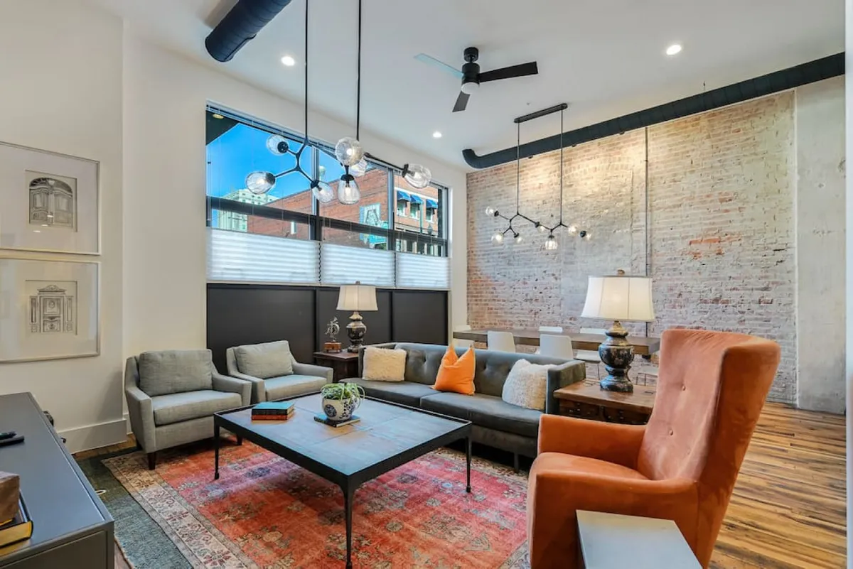 This image showcases a contemporary living area featuring a textured brick wall, large windows that welcome natural light, and a colorful accent chair. Sophisticated lighting fixtures and a well-styled coffee table create a space that feels both stylish and welcoming.