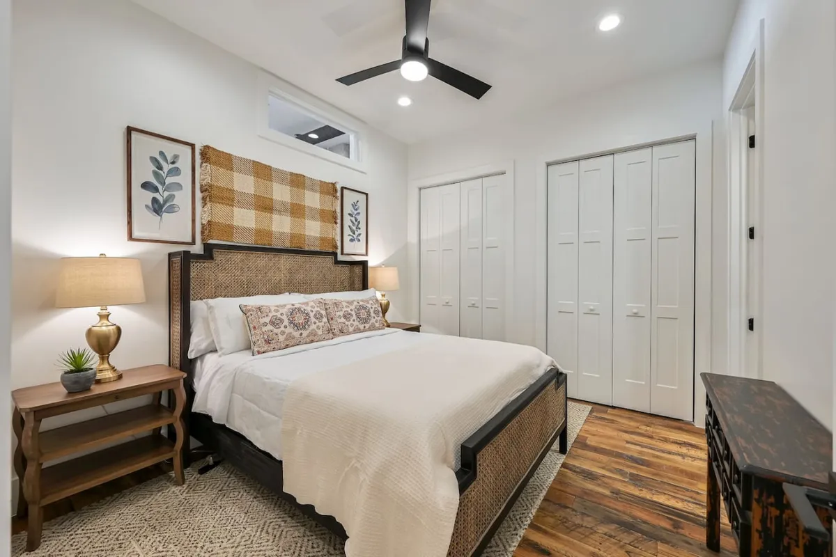 A tranquil bedroom featuring a plush queen bed adorned with decorative pillows, flanked by nightstands with lamps that create a cozy, inviting ambiance. Textured wall art and sunlight filtering through a window enhance the serene atmosphere, highlighting both comfort and style.