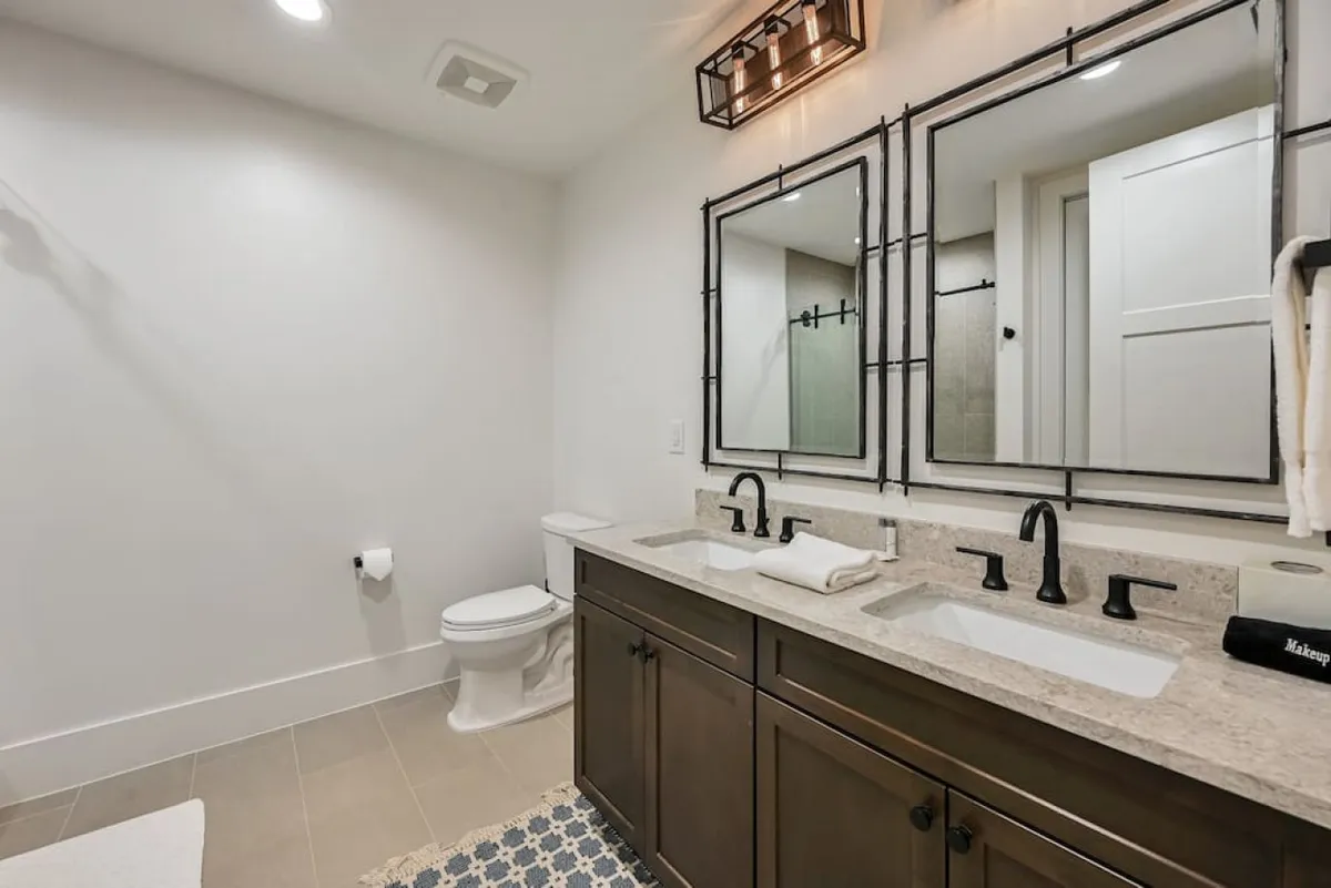 A stylish ensuite bathroom featuring a double vanity with dark wood cabinetry, expansive mirrors, and a modern light fixture. The clean lines and contemporary finishes evoke a sense of calm and sophistication, perfect for unwinding or preparing for the day ahead.